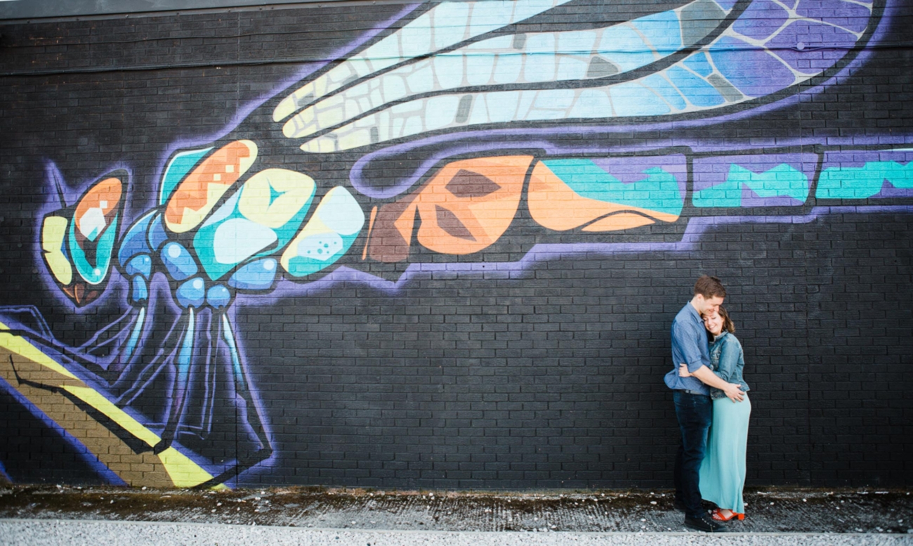 A young couple embrace white stood in front of a graffiti wall depicting a dragonfly