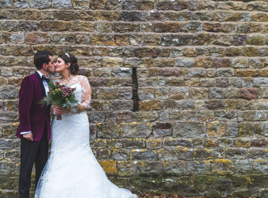 A wedding couple kiss while stood in front of a rustic wall during a winter wedding at The Out Barn in Clough Bottom